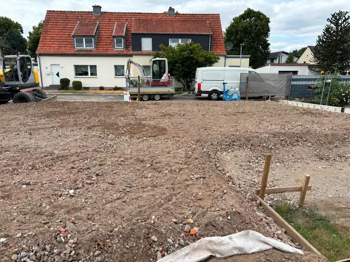 Abgezogenes Baufeld vor einem Wohnhaus, dahinter Minibagger und Anhänger